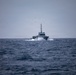 A gray military or coast guard patrol vessel is underway at speed on open, choppy ocean waters under an overcast sky. The vessel is creating a visible bow wave and wake, indicating significant forward momentum. A flock of seabirds is visible in the sky above and behind the vessel.