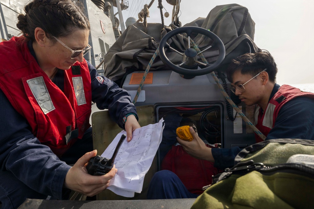 Two personnel wearing red life vests are aboard what appears to be a military or coast guard vessel, reviewing a paper map or chart together. One individual holds a handheld radio while the other holds a yellow device, possibly a GPS unit or emergency beacon. They appear to be conducting navigation or mission planning activities on the deck of the vessel.