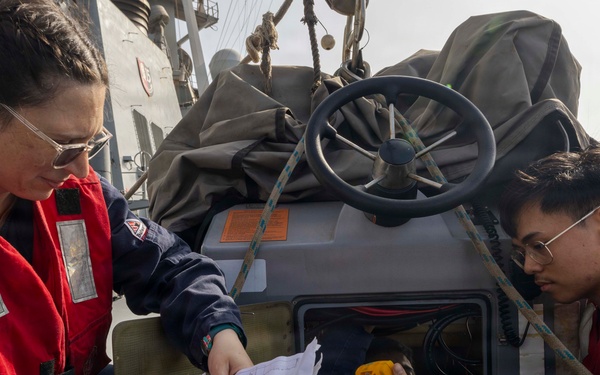 Two personnel wearing red life vests are aboard what appears to be a military or coast guard vessel, reviewing a paper map or chart together. One individual holds a handheld radio while the other holds a yellow device, possibly a GPS unit or emergency beacon. They appear to be conducting navigation or mission planning activities on the deck of the vessel.