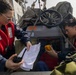 Two personnel wearing red life vests are aboard what appears to be a military or coast guard vessel, reviewing a paper map or chart together. One individual holds a handheld radio while the other holds a yellow device, possibly a GPS unit or emergency beacon. They appear to be conducting navigation or mission planning activities on the deck of the vessel.