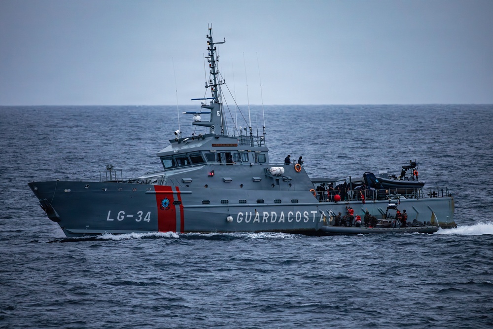A gray coast guard patrol vessel designated LG-34 and marked 'GUARDACOSTAS' is underway on open ocean under overcast skies. The vessel appears to be conducting an active maritime operation, with personnel visible on deck and what appears to be a smaller inflatable craft alongside or attached to the starboard side. The ship features distinctive red diagonal stripes on the bow and is equipped with a tall communications/radar mast.