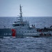 A gray coast guard patrol vessel designated LG-34 and marked 'GUARDACOSTAS' is underway on open ocean under overcast skies. The vessel appears to be conducting an active maritime operation, with personnel visible on deck and what appears to be a smaller inflatable craft alongside or attached to the starboard side. The ship features distinctive red diagonal stripes on the bow and is equipped with a tall communications/radar mast.