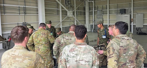 A group of military personnel in various camouflage uniforms are gathered in a circle inside what appears to be a large industrial or hangar-type building. The soldiers appear to be engaged in a briefing, demonstration, or equipment review session. At least two distinct camouflage patterns are visible, suggesting a multinational or joint military exercise.