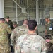 A group of military personnel in various camouflage uniforms are gathered in a circle inside what appears to be a large industrial or hangar-type building. The soldiers appear to be engaged in a briefing, demonstration, or equipment review session. At least two distinct camouflage patterns are visible, suggesting a multinational or joint military exercise.