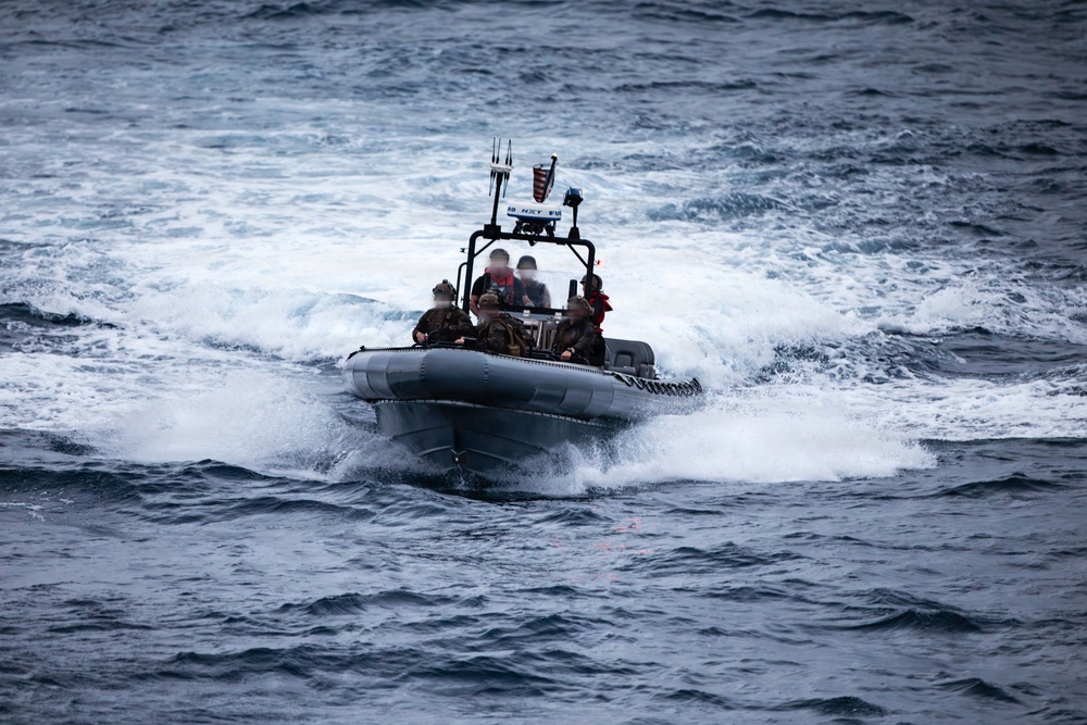 A rigid inflatable boat (RIB) is underway at speed on open ocean waters with choppy conditions. Several personnel are aboard, some wearing military-style camouflage and at least one in a red life vest. The boat appears to be a military or law enforcement vessel equipped with a console mast, antenna array, and what appears to be a small American flag.