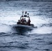 A rigid inflatable boat (RIB) is underway at speed on open ocean waters with choppy conditions. Several personnel are aboard, some wearing military-style camouflage and at least one in a red life vest. The boat appears to be a military or law enforcement vessel equipped with a console mast, antenna array, and what appears to be a small American flag.