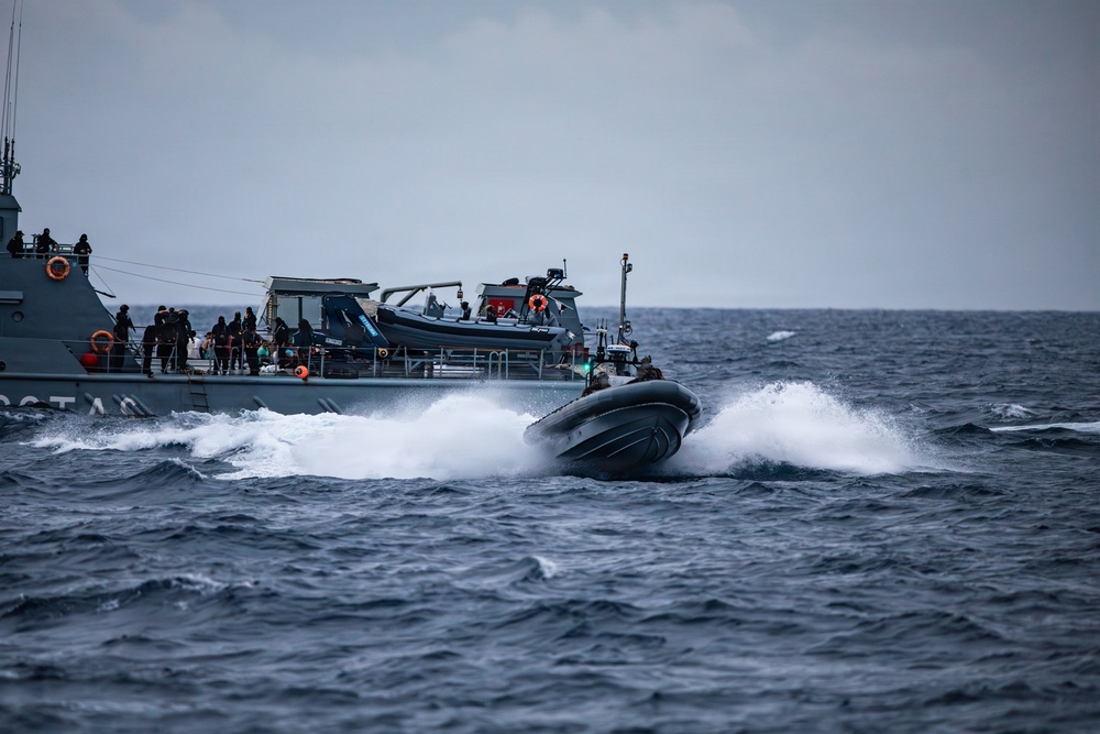 A rigid inflatable boat (RIB) is operating at high speed alongside a larger gray naval or coast guard vessel in open, choppy seas under overcast skies. Personnel are visible on the deck of the larger vessel observing or coordinating with the smaller craft. The scene appears to depict a maritime military or law enforcement exercise or operation.
