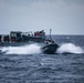 A rigid inflatable boat (RIB) is operating at high speed alongside a larger gray naval or coast guard vessel in open, choppy seas under overcast skies. Personnel are visible on the deck of the larger vessel observing or coordinating with the smaller craft. The scene appears to depict a maritime military or law enforcement exercise or operation.