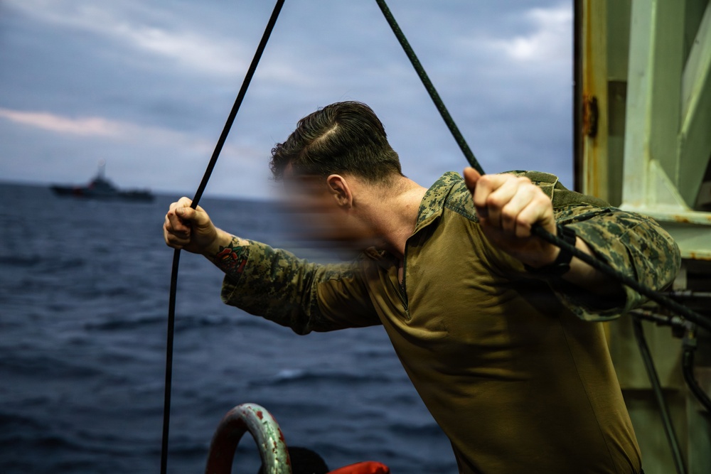 A person wearing military camouflage uniform is gripping ropes or lines on what appears to be the deck of a naval vessel at sea. The image was taken during overcast, low-light conditions with rough-looking open water visible in the background. A second vessel is faintly visible on the horizon in the background.