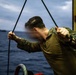 A person wearing military camouflage uniform is gripping ropes or lines on what appears to be the deck of a naval vessel at sea. The image was taken during overcast, low-light conditions with rough-looking open water visible in the background. A second vessel is faintly visible on the horizon in the background.