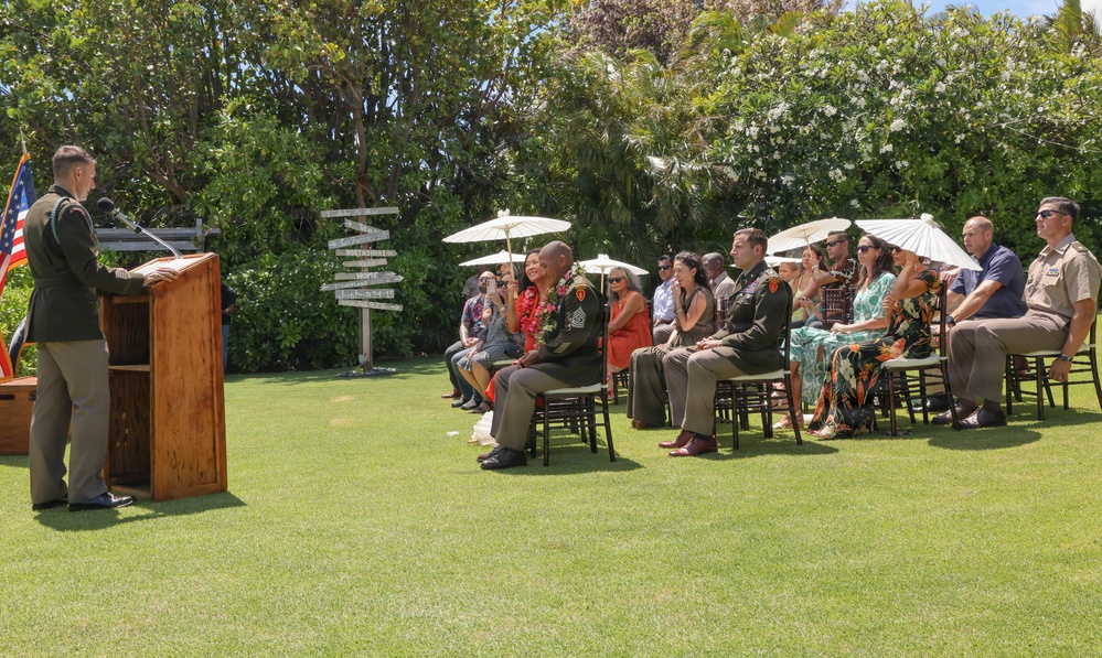 An outdoor military ceremony is taking place on a well-manicured lawn, likely in a tropical location based on the vegetation. A uniformed military officer is speaking at a wooden podium while an audience of military personnel and civilians seated in chairs listens attentively. A senior enlisted service member in the front row wearing a lei suggests this may be a retirement or change of responsibility ceremony.