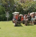 An outdoor military ceremony is taking place on a well-manicured lawn, likely in a tropical location based on the vegetation. A uniformed military officer is speaking at a wooden podium while an audience of military personnel and civilians seated in chairs listens attentively. A senior enlisted service member in the front row wearing a lei suggests this may be a retirement or change of responsibility ceremony.