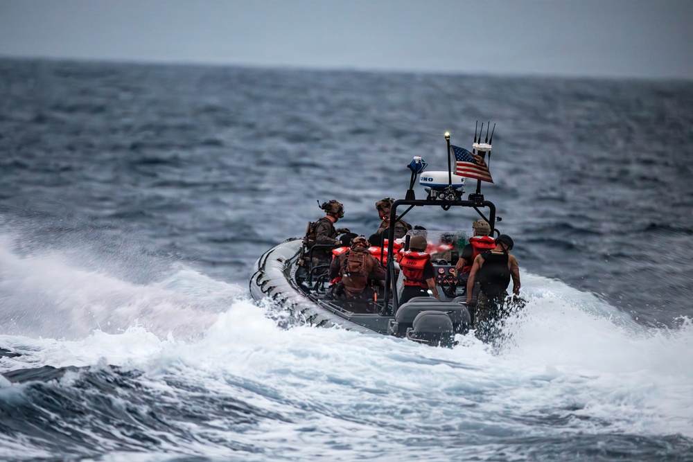 A rigid-hull inflatable boat (RHIB) is underway at high speed through rough open ocean conditions. Multiple personnel are aboard, some wearing military tactical gear and helmets while others wear red life vests. An American flag and what appears to be navigation/communication equipment are mounted on the boat's T-top frame.