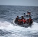 A rigid-hull inflatable boat (RHIB) is underway at high speed through rough open ocean conditions. Multiple personnel are aboard, some wearing military tactical gear and helmets while others wear red life vests. An American flag and what appears to be navigation/communication equipment are mounted on the boat's T-top frame.