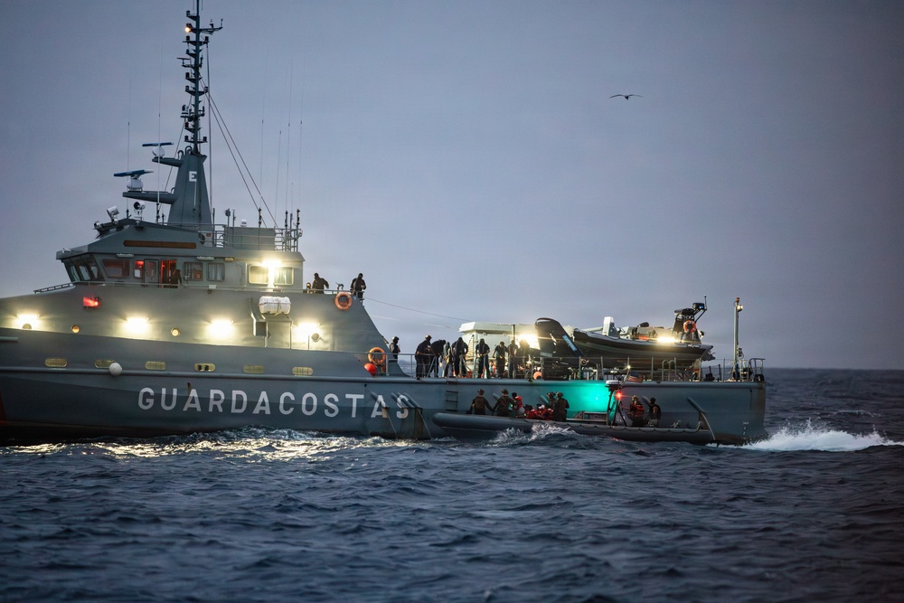 A gray coast guard vessel marked 'GUARDACOSTAS' is operating at sea during dusk or dawn conditions. Multiple personnel are visible on deck and on a smaller rigid inflatable boat (RIB) alongside the main vessel. The scene appears to depict an active maritime operation, possibly a rescue, patrol, or interdiction mission.