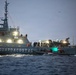 A gray coast guard vessel marked 'GUARDACOSTAS' is operating at sea during dusk or dawn conditions. Multiple personnel are visible on deck and on a smaller rigid inflatable boat (RIB) alongside the main vessel. The scene appears to depict an active maritime operation, possibly a rescue, patrol, or interdiction mission.