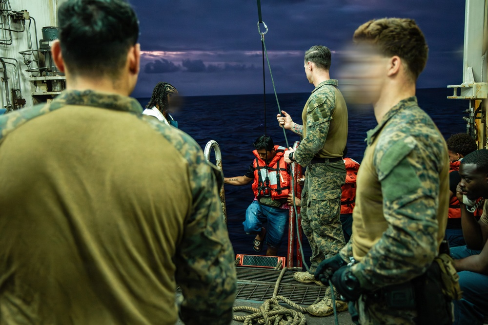 Military personnel in camouflage uniforms are assisting civilians wearing orange life vests onto a vessel at sea during twilight or nighttime conditions. The scene appears to depict a maritime rescue or boarding operation, with service members helping individuals climb aboard from the water or a smaller craft. Several rescued civilians are visible on the deck, some still wearing life vests.
