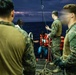 Military personnel in camouflage uniforms are assisting civilians wearing orange life vests onto a vessel at sea during twilight or nighttime conditions. The scene appears to depict a maritime rescue or boarding operation, with service members helping individuals climb aboard from the water or a smaller craft. Several rescued civilians are visible on the deck, some still wearing life vests.