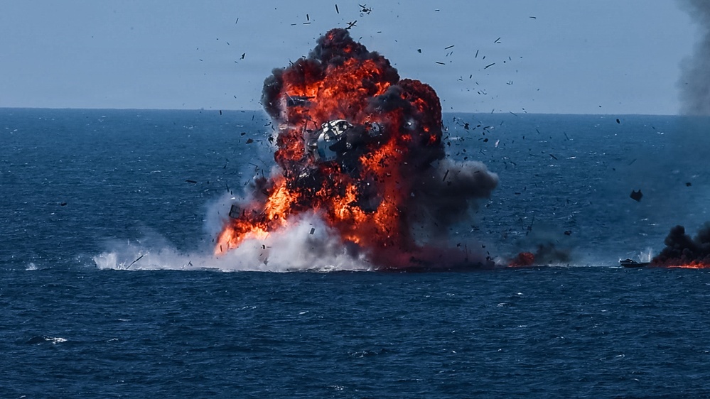 A large explosion is occurring on the surface of an open body of water, producing a massive fireball with red and orange flames accompanied by dense black and grey smoke. Debris is visibly being ejected in multiple directions from the blast epicenter. A secondary fire or smoke source is visible to the right side of the frame.