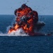 A large explosion is occurring on the surface of an open body of water, producing a massive fireball with red and orange flames accompanied by dense black and grey smoke. Debris is visibly being ejected in multiple directions from the blast epicenter. A secondary fire or smoke source is visible to the right side of the frame.