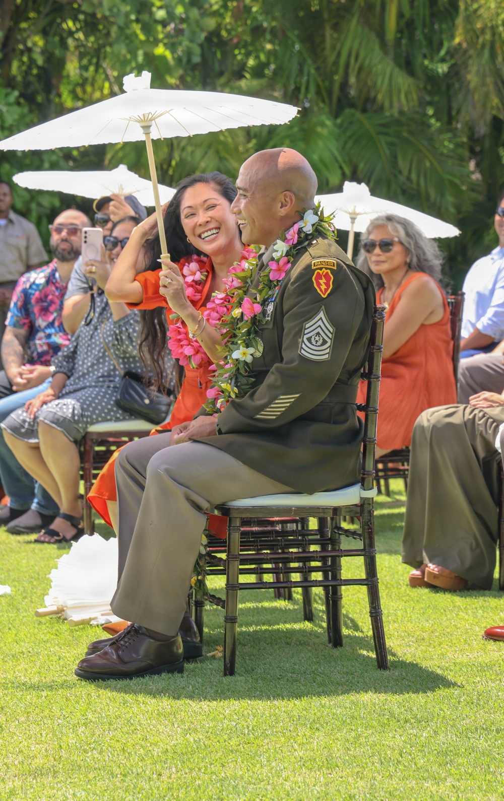 An outdoor ceremony, likely a military retirement or promotion event, taking place on a grass lawn in what appears to be a tropical setting. A soldier in U.S. Army dress green uniform adorned with flower leis sits in a chair while a woman in a red dress holds a white parasol over him and leans in smiling. Guests seated in rows behind them observe the occasion, some holding parasols and one capturing the moment on a smartphone.