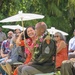 An outdoor ceremony, likely a military retirement or promotion event, taking place on a grass lawn in what appears to be a tropical setting. A soldier in U.S. Army dress green uniform adorned with flower leis sits in a chair while a woman in a red dress holds a white parasol over him and leans in smiling. Guests seated in rows behind them observe the occasion, some holding parasols and one capturing the moment on a smartphone.
