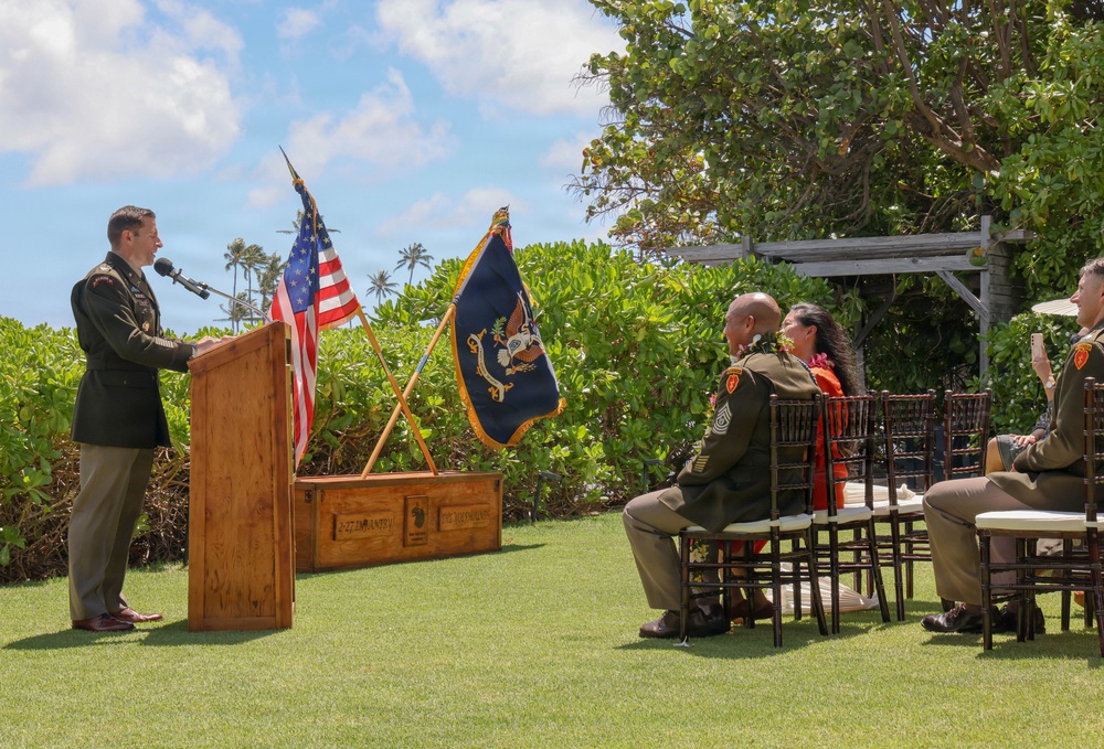 An outdoor military ceremony is taking place on a well-manicured lawn in what appears to be a tropical location, likely Hawaii based on the palm trees and vegetation. A uniformed Army officer is speaking at a wooden podium flanked by the U.S. flag and a dark blue military unit flag. A small audience of military personnel and at least one civilian are seated in chairs facing the speaker.