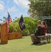 An outdoor military ceremony is taking place on a well-manicured lawn in what appears to be a tropical location, likely Hawaii based on the palm trees and vegetation. A uniformed Army officer is speaking at a wooden podium flanked by the U.S. flag and a dark blue military unit flag. A small audience of military personnel and at least one civilian are seated in chairs facing the speaker.