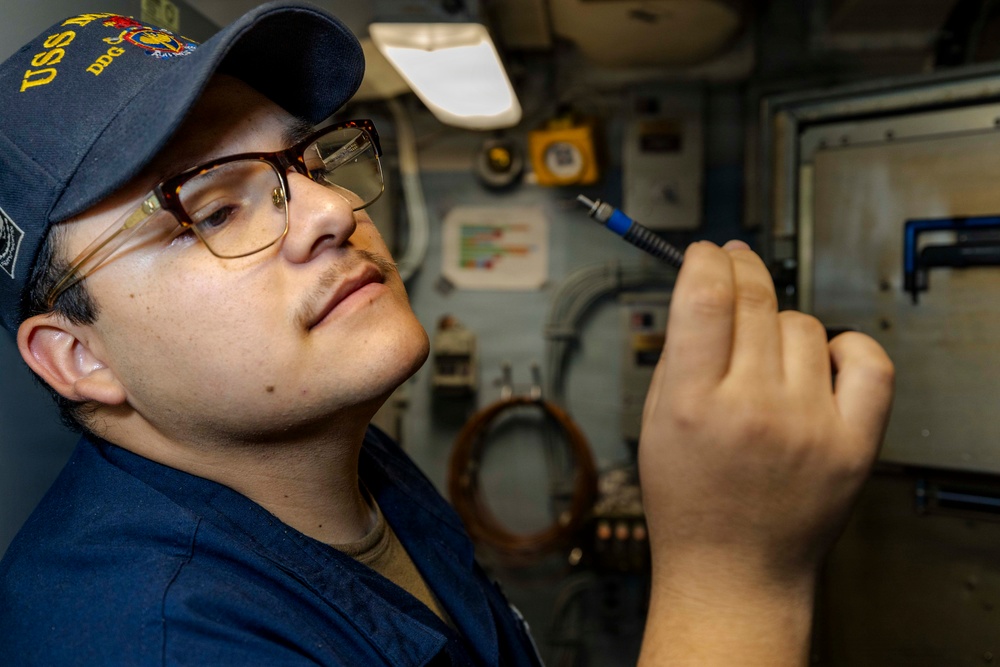 A U.S. Navy sailor wearing a USS ship ball cap (DDG designation visible) and glasses is inspecting or working with a small cable connector or tool inside what appears to be a shipboard compartment. The individual is dressed in Navy working uniform blues and is closely examining the item held in their right hand. The background shows typical naval vessel interior equipment including panels, piping, and electrical components.
