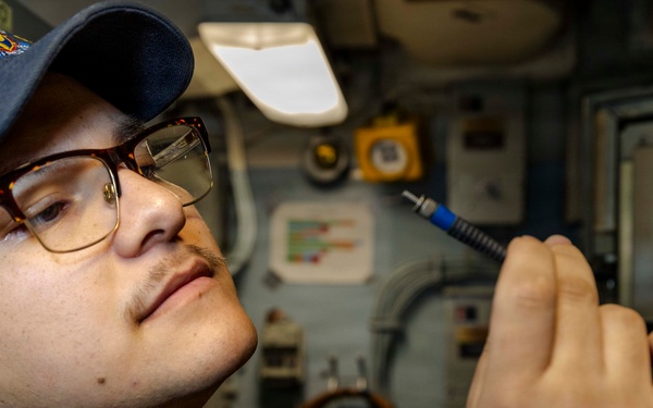 A U.S. Navy sailor wearing a USS ship ball cap (DDG designation visible) and glasses is inspecting or working with a small cable connector or tool inside what appears to be a shipboard compartment. The individual is dressed in Navy working uniform blues and is closely examining the item held in their right hand. The background shows typical naval vessel interior equipment including panels, piping, and electrical components.