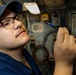 A U.S. Navy sailor wearing a USS ship ball cap (DDG designation visible) and glasses is inspecting or working with a small cable connector or tool inside what appears to be a shipboard compartment. The individual is dressed in Navy working uniform blues and is closely examining the item held in their right hand. The background shows typical naval vessel interior equipment including panels, piping, and electrical components.
