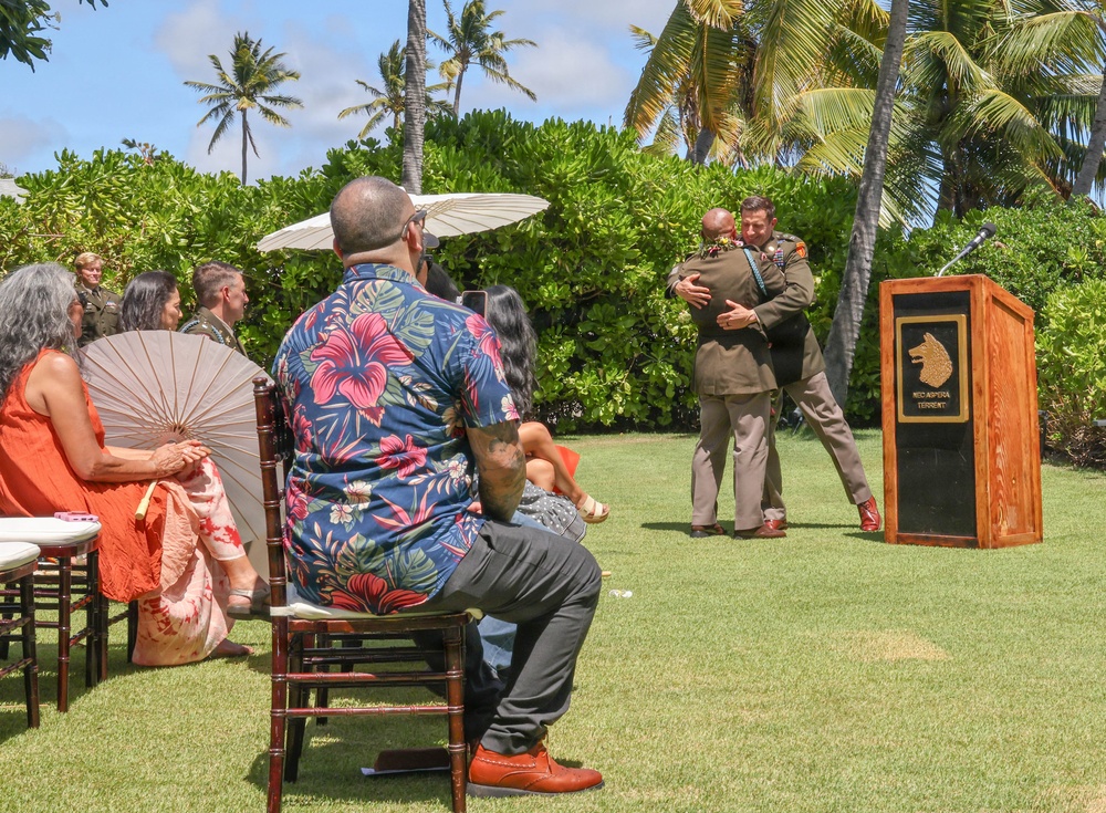 An outdoor military or official ceremony is taking place on a well-manicured lawn in what appears to be a tropical location, likely Hawaii, based on the palm trees and Hawaiian-print attire worn by attendees. Two uniformed individuals are embracing near a wooden podium, suggesting a moment of congratulation or recognition. Seated guests, some holding parasols for shade, are watching the proceedings.