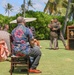 An outdoor military or official ceremony is taking place on a well-manicured lawn in what appears to be a tropical location, likely Hawaii, based on the palm trees and Hawaiian-print attire worn by attendees. Two uniformed individuals are embracing near a wooden podium, suggesting a moment of congratulation or recognition. Seated guests, some holding parasols for shade, are watching the proceedings.