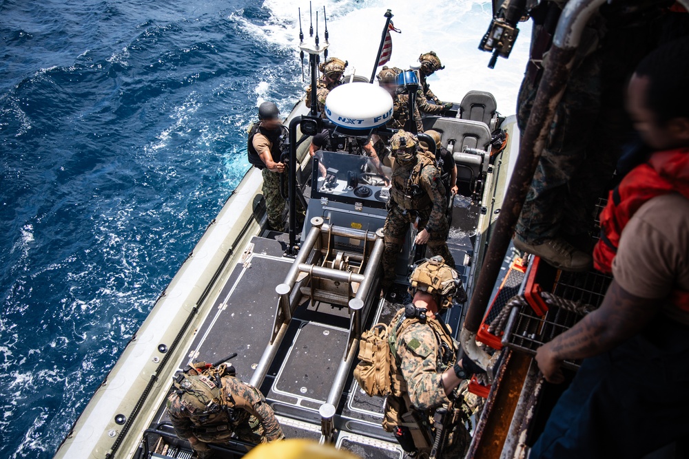 The image shows military personnel, appearing to be U.S. Marines and Navy sailors, conducting a maritime boarding or transfer operation from a rigid-hull inflatable boat (RHIB). Personnel are equipped with full combat gear, tactical vests, helmets, and weapons. An American flag is visible on the vessel, and the operation appears to be taking place in open ocean conditions.