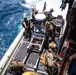 The image shows military personnel, appearing to be U.S. Marines and Navy sailors, conducting a maritime boarding or transfer operation from a rigid-hull inflatable boat (RHIB). Personnel are equipped with full combat gear, tactical vests, helmets, and weapons. An American flag is visible on the vessel, and the operation appears to be taking place in open ocean conditions.