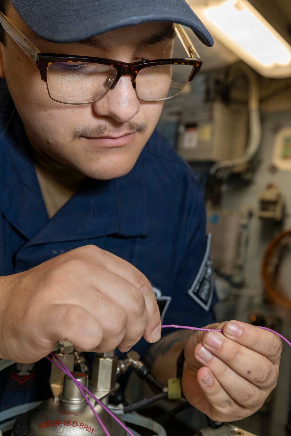 A military service member wearing a blue uniform and cap is working on a mechanical or electrical component, handling a purple/pink wire connected to a metallic fitting or valve assembly. The setting appears to be the interior of a naval vessel, indicated by the confined space, piping, and equipment visible in the background. The individual is focused on the task, suggesting maintenance or repair work.