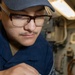 A military service member wearing a blue uniform and cap is working on a mechanical or electrical component, handling a purple/pink wire connected to a metallic fitting or valve assembly. The setting appears to be the interior of a naval vessel, indicated by the confined space, piping, and equipment visible in the background. The individual is focused on the task, suggesting maintenance or repair work.