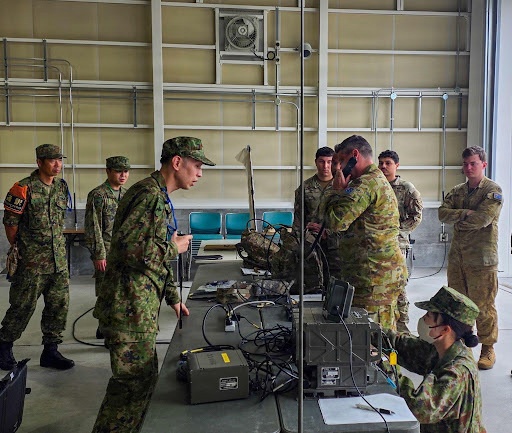 Military personnel from what appear to be two different nations are gathered in an indoor facility around a table with communications and electronic equipment. Soldiers in Japanese Ground Self-Defense Force (JGSDF) uniforms are interacting with soldiers wearing Australian Defence Force (ADF) multicam uniforms. The scene appears to depict a joint military training or equipment demonstration exercise.