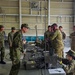 Military personnel from what appear to be two different nations are gathered in an indoor facility around a table with communications and electronic equipment. Soldiers in Japanese Ground Self-Defense Force (JGSDF) uniforms are interacting with soldiers wearing Australian Defence Force (ADF) multicam uniforms. The scene appears to depict a joint military training or equipment demonstration exercise.