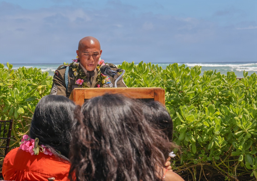 A military servicemember in uniform is speaking at a wooden podium during an outdoor ceremony near a beach. The speaker is wearing a lei made of flowers and leaves, along with military medals and insignia on their uniform. Two audience members with dark hair are visible in the foreground, with lush green vegetation and ocean waves visible in the background.