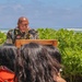 A military servicemember in uniform is speaking at a wooden podium during an outdoor ceremony near a beach. The speaker is wearing a lei made of flowers and leaves, along with military medals and insignia on their uniform. Two audience members with dark hair are visible in the foreground, with lush green vegetation and ocean waves visible in the background.