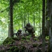 Two soldiers in full camouflage combat gear are positioned on a moss-covered rocky outcrop in a dense deciduous forest. They appear to be in a tactical defensive or observation posture, with weapons raised and aimed forward. The lush green foliage and dappled lighting suggest a temperate forest environment, likely during a military training exercise.