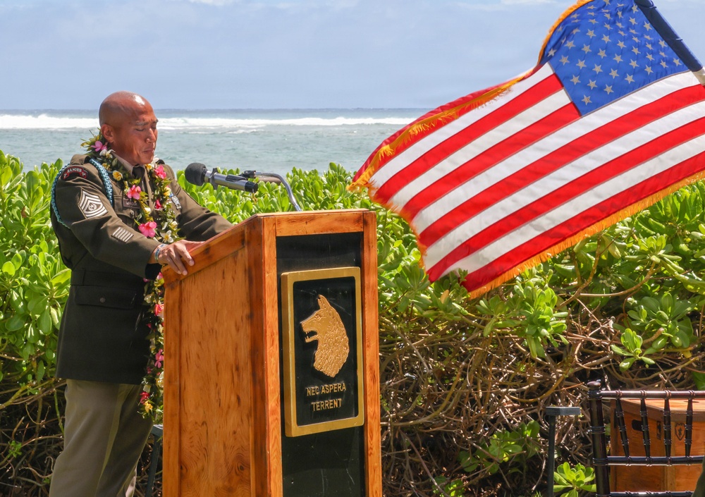 A military official in dress uniform adorned with flower leis speaks at a wooden podium during an outdoor ceremony near the ocean. The podium bears a wolf/dog emblem and the Latin motto 'NEC ASPERA TERRENT' (Nothing Deters Us). An American flag with gold fringe flies prominently to the right, suggesting a formal military or commemorative event, likely in Hawaii.