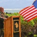A military official in dress uniform adorned with flower leis speaks at a wooden podium during an outdoor ceremony near the ocean. The podium bears a wolf/dog emblem and the Latin motto 'NEC ASPERA TERRENT' (Nothing Deters Us). An American flag with gold fringe flies prominently to the right, suggesting a formal military or commemorative event, likely in Hawaii.