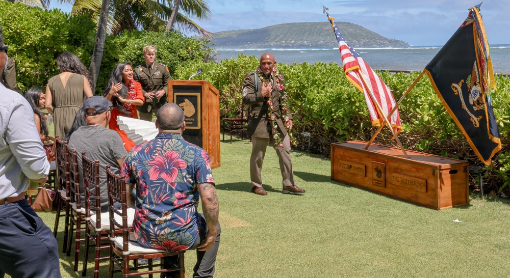 An outdoor military ceremony is taking place on a grassy lawn near the ocean, likely in Hawaii based on the tropical vegetation, palm trees, and mountainous coastline in the background. A man in military dress uniform adorned with a floral lei stands at the center, appearing to be the honoree of a promotion or retirement ceremony. Guests are seated in rows of wooden chairs while flags — including the American flag and what appears to be a unit colors flag — are displayed alongside a wooden ceremonial chest.