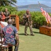 An outdoor military ceremony is taking place on a grassy lawn near the ocean, likely in Hawaii based on the tropical vegetation, palm trees, and mountainous coastline in the background. A man in military dress uniform adorned with a floral lei stands at the center, appearing to be the honoree of a promotion or retirement ceremony. Guests are seated in rows of wooden chairs while flags — including the American flag and what appears to be a unit colors flag — are displayed alongside a wooden ceremonial chest.