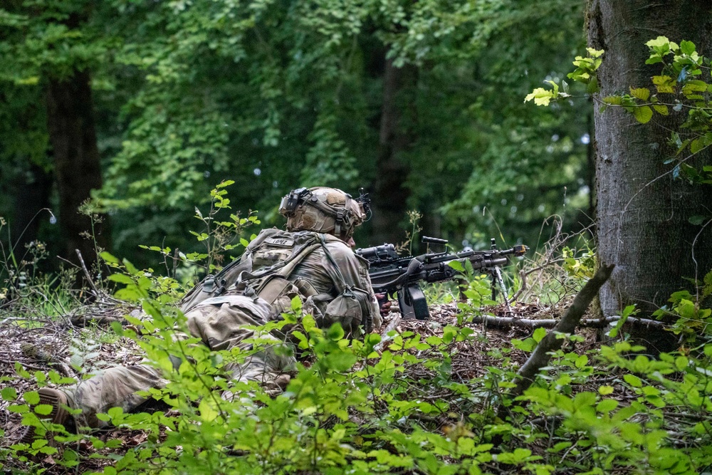 A soldier in full combat gear is positioned prone in a wooded area, aiming a machine gun supported by a bipod. The soldier is wearing MultiCam camouflage uniform, a tactical helmet with mounted accessories, and a full rucksack. The setting appears to be a training exercise or tactical operation in a dense forest environment.