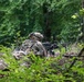 A soldier in full combat gear is positioned prone in a wooded area, aiming a machine gun supported by a bipod. The soldier is wearing MultiCam camouflage uniform, a tactical helmet with mounted accessories, and a full rucksack. The setting appears to be a training exercise or tactical operation in a dense forest environment.