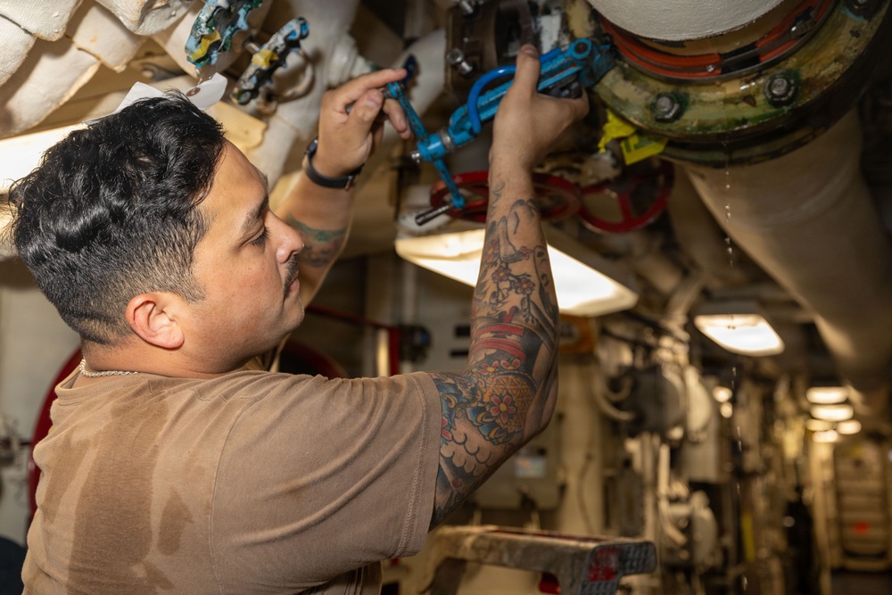 A tattooed male technician or maintenance worker is working on overhead piping and valve systems in what appears to be a ship's engine room or mechanical compartment. He is wearing a tan/brown t-shirt and a wristwatch, and is manipulating blue-handled valves on a complex pipe assembly. The environment is a narrow industrial corridor with multiple pipes, fittings, and mechanical components visible.