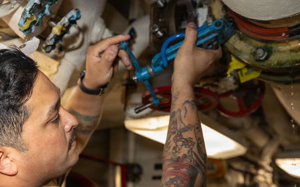 A tattooed male technician or maintenance worker is working on overhead piping and valve systems in what appears to be a ship's engine room or mechanical compartment. He is wearing a tan/brown t-shirt and a wristwatch, and is manipulating blue-handled valves on a complex pipe assembly. The environment is a narrow industrial corridor with multiple pipes, fittings, and mechanical components visible.